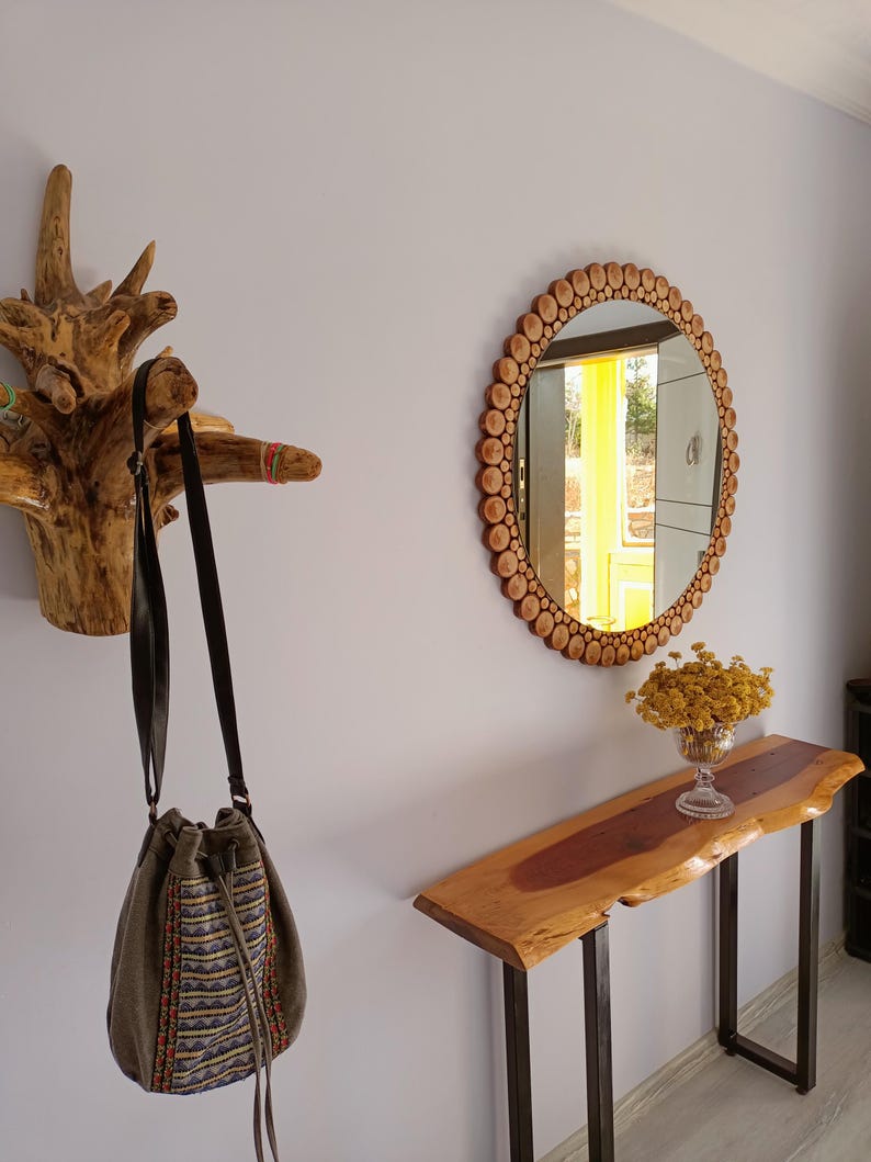 May include: A rustic entryway scene featuring a wooden coat rack, a round mirror with a wooden frame, and a live-edge console table. A green and patterned drawstring bag hangs from the coat rack. A vase of yellow flowers sits on the table.