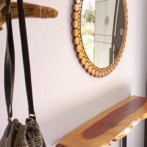 May include: A round mirror with a wooden frame made of small, circular wood pieces. A wooden console table with a live edge is below the mirror. A canvas bag with a strap hangs from a wooden branch.
