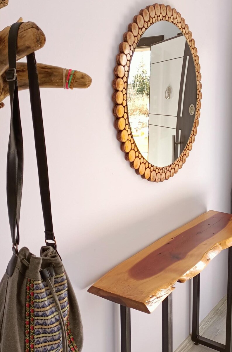 May include: A round mirror with a wooden frame made of small, circular wood pieces. A wooden console table with a live edge is below the mirror. A canvas bag with a strap hangs from a wooden branch.