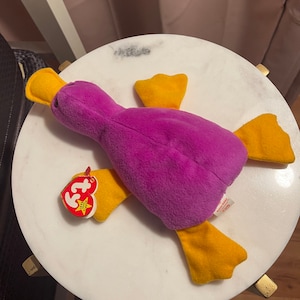 May include: Purple and yellow plush toy duck with a red and white "Ty" tag. The tag has a white star and the text "Beanie Babies".