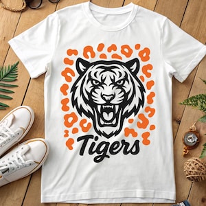 Puede incluir: Camiseta blanca con un diseño de cabeza de tigre en blanco y negro, manchas de leopardo naranja y la palabra "Tigers". La camiseta está sobre una superficie de madera. Se ven zapatillas blancas.