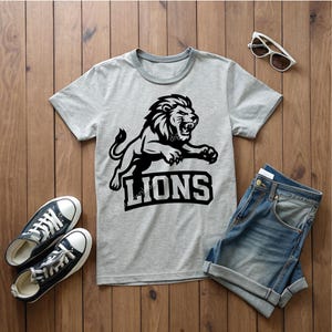 May include: A grey t-shirt featuring a black lion design and the word "LIONS". The image includes blue denim shorts, black trainers, and sunglasses, all arranged on a wooden surface.