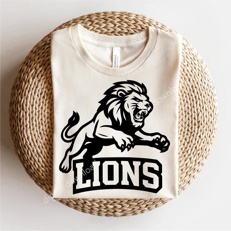 May include: Cream-coloured t-shirt with a black graphic of a roaring lion in mid-leap. The word "LIONS" is printed in a bold, black, block font below the lion. The shirt is made of a soft material.
