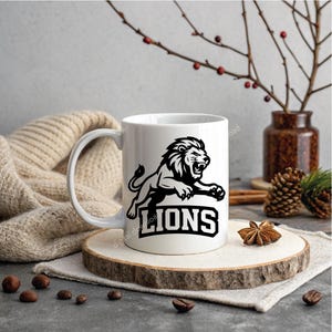 May include: White ceramic mug with a black and white lion illustration and the word "LIONS". The mug is presented on a wooden slice, with a neutral backdrop and decorative items, including a vase and pine cones.