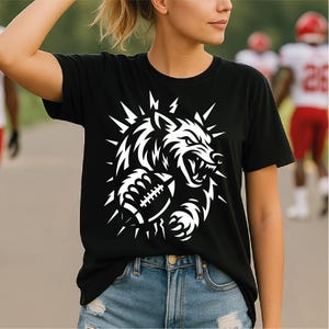 Puede incluir: Camiseta negra con un diseño gráfico blanco de un lobo gruñendo que sostiene un balón de fútbol americano. El diseño del lobo está rodeado de elementos puntiagudos. La camiseta se combina con vaqueros azules.