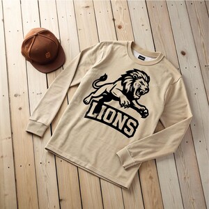 May include: A beige long-sleeved sweatshirt featuring a black lion graphic and the word "LIONS". A brown baseball cap with a curved peak is next to the sweatshirt. The sweatshirt is made of a soft material.