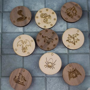 Puede incluir: Una colección de fichas de juego de madera con varios diseños grabados, incluyendo una serpiente, una abeja, un caballo, una araña y un murciélago. Las fichas están dispuestas sobre una superficie embaldosada, con dados visibles en el fondo. Las fichas miden unos 5 cm de diámetro.