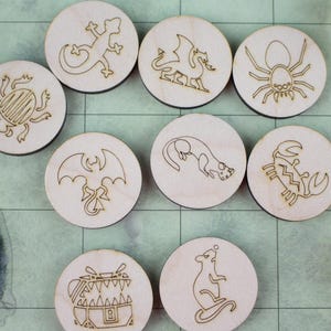 Könnte beinhalten: Zehn Holz-Token mit lasergravierten Designs von Fantasy-Kreaturen, darunter eine Eidechse, ein Drache und eine Spinne. Die Token sind rund und hell, mit dunklen Umrissen. Würfel sind im Hintergrund zu sehen, was auf ein Tabletop-Spiel hindeutet.