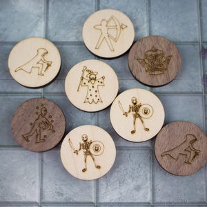 Puede incluir: Fichas de juego de madera con diseños grabados de personajes de fantasía. Las fichas redondas, algunas claras y otras oscuras, representan un mago, un arquero y un guerrero esqueleto. Las fichas están dispuestas sobre una superficie embaldosada.