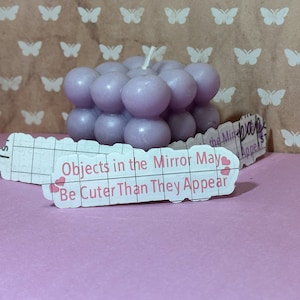 Könnte beinhalten: Eine lavendelfarbene Blasenkerze mit dem Text "Objects in the Mirror May Be Cuter Than They Appear" auf einem weißen Etikett mit rosa Herzen.