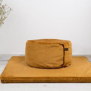 Puede incluir: Un cojín de meditación amarillo mostaza y un zabuton a juego, un cojín de suelo japonés tradicional. El cojín tiene una cremallera y una etiqueta negra con la palabra "Zafu" en él.