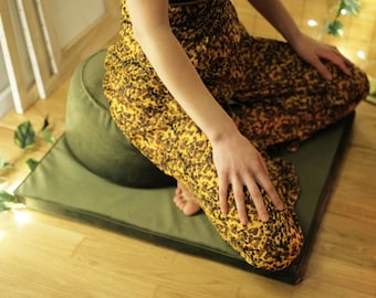 Puede incluir: Una persona sentada en un cojín de meditación verde con una falda de estampado amarillo y marrón. El cojín está sobre un suelo de madera.