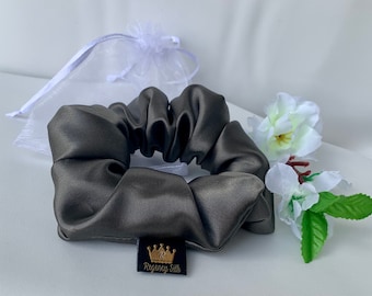 May include: A dark gray satin scrunchie with a small black label that reads "Regency Silk" with a gold crown logo. The scrunchie is next to a white organza gift bag and white flowers with green leaves.