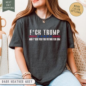 May include: Dark heather grey t-shirt with the text "F*CK TRUMP" above the text "AND F*CK YOU FOR VOTING FOR HIM". The text is in white, with a red and blue stripe design. The shirt is a crew neck.