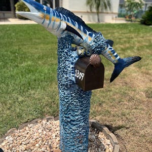 May include: A blue and white marlin sculpture atop a textured blue post, serving as a mailbox. The mailbox is brown with the number 1309 in white. A seashell and a turtle are also part of the design.