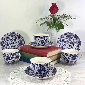 May include: Four blue and white floral teacups and saucers with a stack of books in the background. The teacups have a delicate floral pattern and are accented with a gold rim.