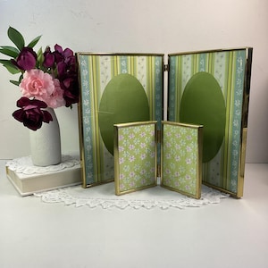 May include: A collection of decorative photo frames with gold-coloured trim. The frames have a green and white striped background with an oval cutout. Two smaller frames display a floral pattern. A vase of flowers is on the left.