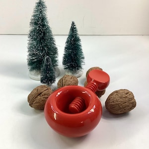 May include: A red nutcracker with a screw mechanism, walnuts, and miniature green Christmas trees with white bases. The nutcracker is a bright, glossy red.