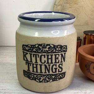 May include: A cream-coloured ceramic kitchen storage jar with a white top and a blue rim. The words "KITCHEN THINGS" are printed in black on the front of the jar.