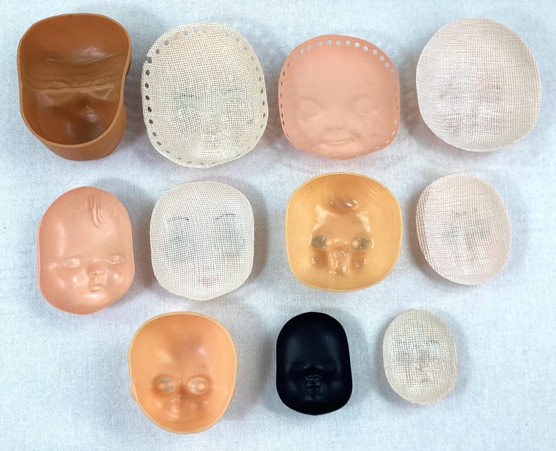May include: Assortment of ten doll head molds, including a brown plastic mold, a black plastic mold, and eight white plastic molds. Some of the molds have a mesh texture.