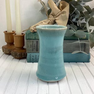 May include: A turquoise ceramic vase with a flared top and rounded base. The vase is displayed on a white wooden surface, with vintage books and candles in the background. The vase has a smooth, glossy finish.