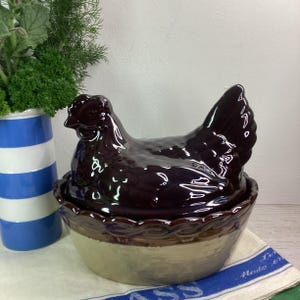 May include: A dark brown ceramic chicken-shaped lid sits atop a round, beige-coloured bowl with a scalloped rim. The chicken figure features a glossy finish and detailed moulded feathers, while the bowl has a smooth, light-coloured base.