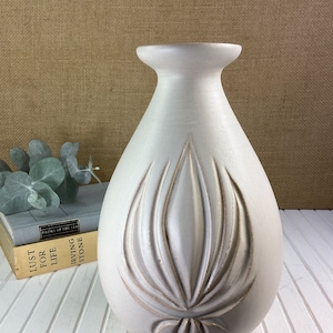 May include: A white ceramic vase with a carved leaf design. The vase has a rounded body and a narrow neck.