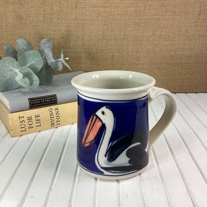 May include: A white ceramic mug featuring a blue and white pelican design. The pelican has a pink beak and black eyes. The mug has a white handle.