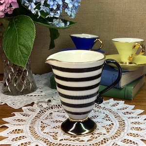 May include: A black and white striped ceramic creamer with a gold rim and a black base. The creamer is sitting on a white lace doily.