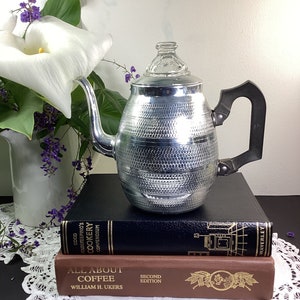 May include: A silver percolator coffee pot with a glass lid and a black handle sits on top of two books. The top book is black with gold lettering that reads "Good Housekeeping's Cookery Compendium". The bottom book is brown with gold lettering that reads "All About Coffee, Second Edition, William H. Ukers".