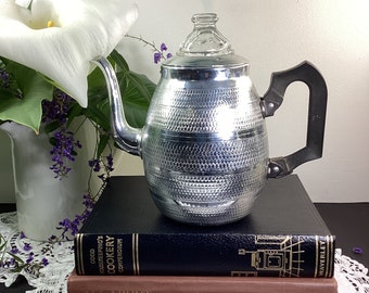 Op de afbeelding: Een zilveren percolator koffiepot met een glazen deksel en een zwarte handgreep staat op twee boeken. Het bovenste boek is zwart met gouden letters die "Good Housekeeping's Cookery Compendium" lezen. Het onderste boek is bruin met gouden letters die "All About Coffee, Second Edition, William H. Ukers" lezen.