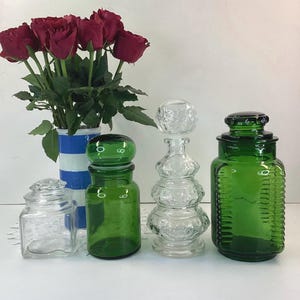 May include: A collection of glass jars and bottles in various shapes and colours, including clear and green. A bouquet of red roses sits in a blue and white striped vase. The items are arranged on a white surface, creating a still life composition.