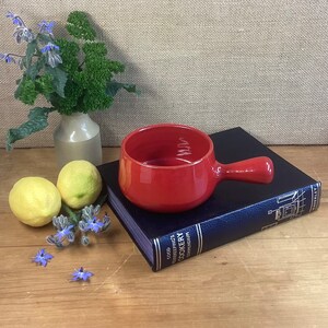 May include: A red ceramic ramekin with a handle. The ramekin is sitting on top of a blue book titled "Good Housekeeping's Cookery Compendium".