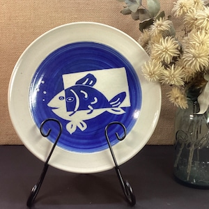 May include: A white ceramic plate with a blue and white fish design. The plate is on a black stand.