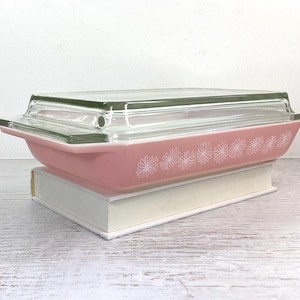 May include: A pink glass baking dish with a clear glass lid. The dish has a white floral pattern around the edge. The dish is sitting on a white surface.