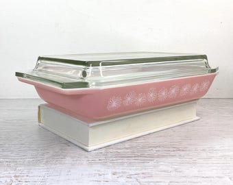 Può includere: Una pirofila di vetro rosa con un coperchio di vetro trasparente. La pirofila ha un motivo floreale bianco attorno al bordo. La pirofila è appoggiata su una superficie bianca.