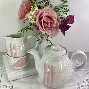 May include: A pink and white teapot and creamer set with a polka dot and stripe design. The teapot has a lid and a handle. The creamer has a spout and a handle. The set is sitting on a white book with the words "The Book of" on the cover.