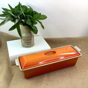 May include: An orange rectangular ceramic baking dish with a lid, featuring a handle and white trim. A small vase with green foliage and the text "Maille Moutarde à l'ancienne" is in the background.