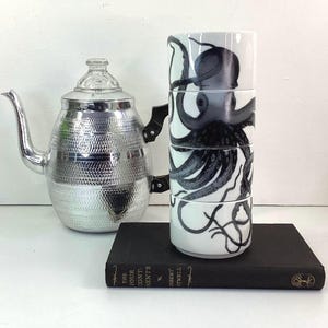 May include: A silver teapot with a textured body and a glass lid sits beside a stack of four white cups. Each cup features a black octopus design. A black book is in the foreground.