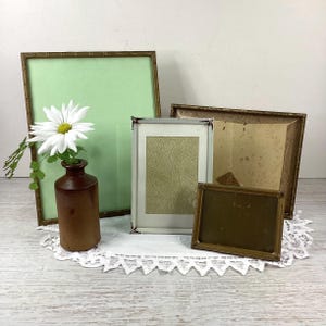 May include: A collection of antique picture frames in different shapes and sizes. A brown ceramic vase holds a white daisy. The frames include a bamboo-style frame with a pale green backing, and a small brass frame.