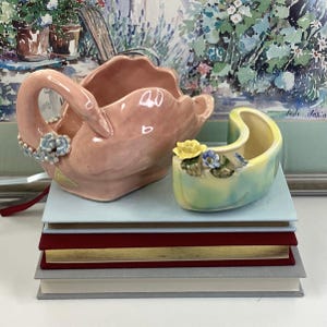May include: Two ceramic planters, one shaped like a swan in pink with blue flowers and the other a crescent shape in yellow, green, and blue with flowers. The planters are stacked on top of four books.