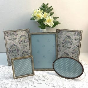 May include: A selection of antique-style picture frames in different shapes and sizes. The frames have decorative gold and silver-toned edges. Some frames display floral patterned backgrounds, while others have a light blue dotted design. A vase of flowers is in the background.