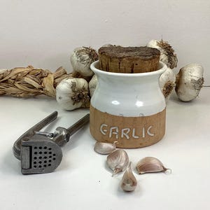 May include: White ceramic garlic jar with a cork lid and the word "GARLIC" written on the front. The jar is surrounded by garlic bulbs and a garlic press.