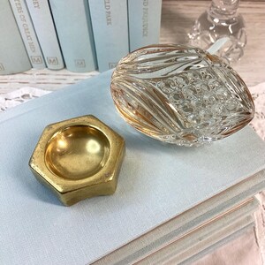 May include: A clear, cut-glass decorative piece with a gold rim and a small, hexagonal brass dish. The glass piece has a textured, oval shape. The brass dish is small and has a shallow bowl. The items are on a stack of light blue books.