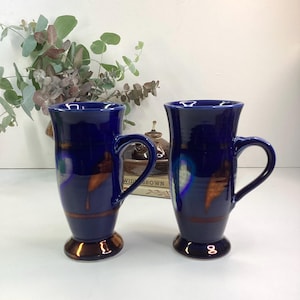 May include: Two tall, cobalt blue ceramic mugs with a flared top and a rounded handle. The mugs have a brown base and a brown band around the middle. The mugs are decorated with streaks of brown and blue. The mugs are suitable for coffee or tea.