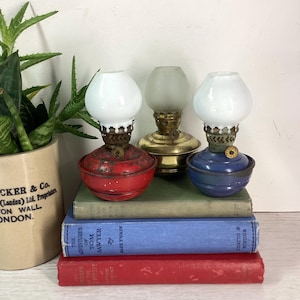 May include: Three vintage oil lamps with glass shades, in red, brass, and blue. They are placed on a stack of old books, including "The Adventures of Tom Sawyer" by Mark Twain. A potted plant is in the background.