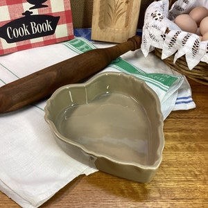 May include: A heart-shaped ceramic baking dish with a light brown glaze. The dish is sitting on a white kitchen towel.