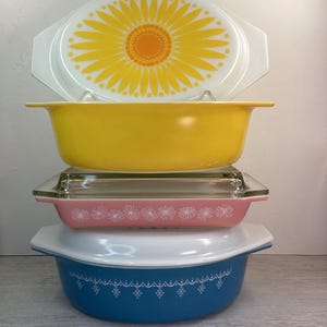 May include: A collection of vintage Pyrex casserole dishes in a stack. The top dish has a white lid with a yellow and orange sunflower design. Beneath is a yellow dish, a pink dish with white floral accents, and a blue dish with white decorative patterns.