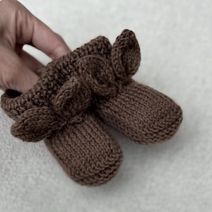 May include: A pair of brown knitted baby booties. The booties are a solid brown color and feature a ribbed cuff and a decorative leaf-like design on the front. The soft, textured knit suggests a cozy and comfortable feel.