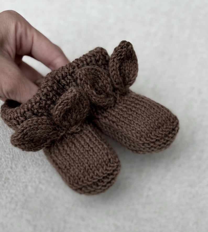 May include: A pair of brown knitted baby booties. The booties are a solid brown color and feature a ribbed cuff and a decorative leaf-like design on the front. The soft, textured knit suggests a cozy and comfortable feel.